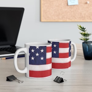 May include: Two white ceramic mugs with a design of the American flag. The flag is faded and has a vintage look. The mugs are on a white surface.