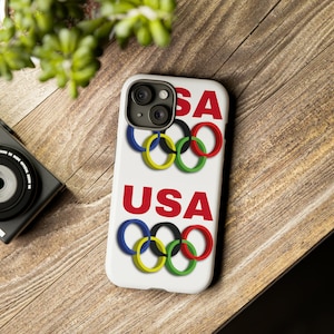 May include: White phone case with a red, white, and blue USA design and Olympic rings. The case has a camera lens cutout.
