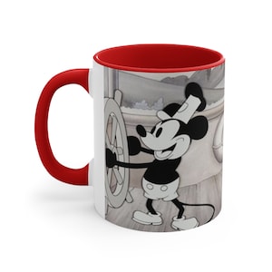 Könnte beinhalten: Eine weiße Keramiktasse mit rotem Rand und Henkel. Die Tasse zeigt eine Schwarzweißillustration von Mickey Mouse, der als Seemann verkleidet ist und ein Steuerrad hält.