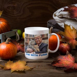 Könnte beinhalten: Weiße Keramiktasse mit einem Bild eines Mannes, der zwei Katzen hält, mit dem Text "I'M WITH PURR". Die Tasse ist von Kürbissen, Äpfeln und Herbstblättern umgeben.