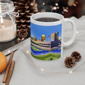 Könnte beinhalten: Weiße Keramiktasse mit einer bunten Illustration eines Baseballstadions mit einer Skyline im Hintergrund. Die Tasse ist mit Kaffee gefüllt.