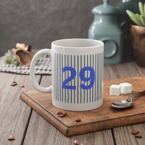 Könnte beinhalten: Weiße Keramiktasse mit einer blauen Zahl 29 darauf gedruckt. Die Tasse hat ein schwarz-weiß gestreiftes Muster.