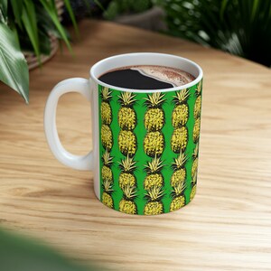Könnte beinhalten: Weiße Keramiktasse mit grünem Hintergrund und einem sich wiederholenden Muster aus gelben Ananas. Die Tasse ist mit Kaffee gefüllt.