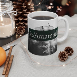 Op de afbeelding: Witte keramische mok met een zwart-witte afbeelding van een gebouw en wolken. De tekst "//Amaranth" is in groen op de mok gedrukt.