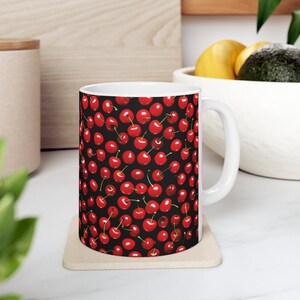 Könnte beinhalten: Weiße Keramiktasse mit schwarzem Hintergrund und einem sich wiederholenden Muster aus roten Kirschen. Die Tasse hat einen weißen Henkel.