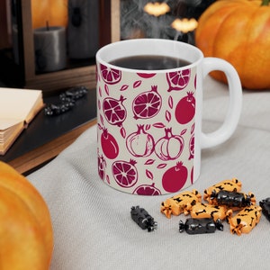 Könnte beinhalten: Weiße Keramiktasse mit einem Muster aus roten Granatäpfeln, Orangen und Kürbissen. Die Tasse ist mit Kaffee gefüllt und hat einen weißen Henkel.