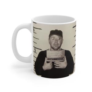 Może przedstawiać: Biały kubek ceramiczny z czarno-białym zdjęciem mężczyzny w policyjnym zdjęciu. Mężczyzna ma na sobie czapkę z daszkiem i trzyma znak z napisem "Sheriff County Jail, Harrison, MI".