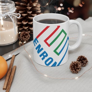 May include: White ceramic mug with a blue, red, and green geometric design and the word "ENRON" printed on the side. The mug is filled with coffee and is sitting on a white surface with string lights.