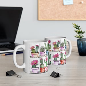 Könnte beinhalten: Weiße Keramiktasse mit einem bunten Kaktusmuster. Die Tasse zeigt verschiedene Kakteen in verschiedenen Töpfen, mit rosa Blüten und grünen Blättern.