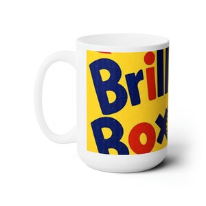 Könnte beinhalten: Weiße Keramiktasse mit gelbem Hintergrund und blauem und rotem Text, der "Brillo Box" lautet.