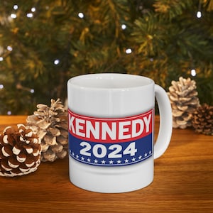 Op de afbeelding: Witte keramische koffiemok met een blauw, rood en wit ontwerp. Het ontwerp bevat de tekst "KENNEDY 2024" met sterren rond de tekst.