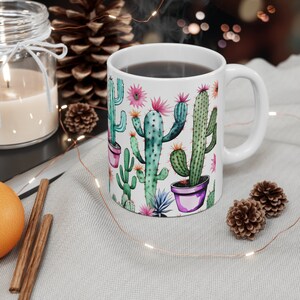 Könnte beinhalten: Weiße Keramiktasse mit einem bunten Kaktusdruck. Die Tasse hat einen Henkel und ist mit einer dunklen Flüssigkeit gefüllt.