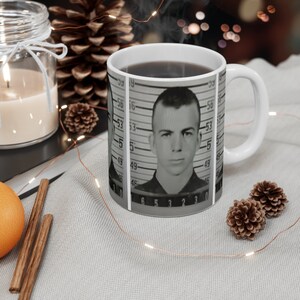 Könnte beinhalten: Weiße Keramiktasse mit einem schwarz-weißen Mugshot eines Mannes mit dem Text "5 6 3 2 1 0" und "5 6 3 2 1 0" auf der Tasse.