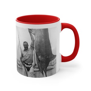 Peut inclure: Une photo en noir et blanc d'une personne tenant un gros marlin. La personne porte une chemise boutonnée et une ceinture. Le poisson est suspendu au bras de la personne. La photo est imprimée sur une tasse en céramique blanche avec un bord rouge.