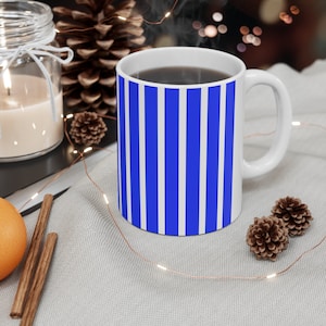 Könnte beinhalten: Eine weiße Keramiktasse mit einem blau-weiß gestreiften Muster. Die Tasse ist mit einer dunklen Flüssigkeit gefüllt und Dampf steigt aus der Oberseite auf. Die Tasse steht auf einer weißen Oberfläche mit einer Lichterkette und Tannenzapfen.