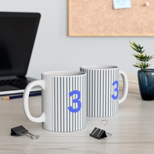 Könnte beinhalten: Zwei weiße Keramikbecher mit blauen Streifen und der Zahl "3" darauf gedruckt. Die Becher stehen auf einer hellen Holzoberfläche.