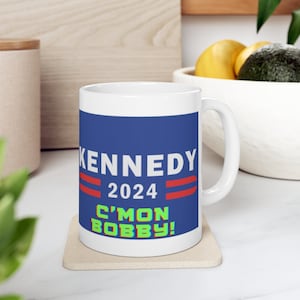 Puede incluir: Taza de café de cerámica blanca con fondo azul y el texto "KENNEDY 2024 C'MON BOBBY!" en blanco, rojo y verde.