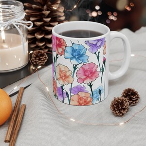 Könnte beinhalten: Weiße Keramiktasse mit einem bunten Blumendesign mit rosa, blauen und orangefarbenen Nelken. Die Tasse ist mit einer dunklen Flüssigkeit gefüllt und hat einen weißen Henkel.