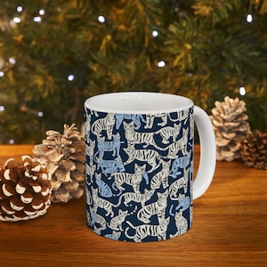 Peut inclure: Une tasse en céramique blanche avec un fond bleu et un motif répété de chats rayés blancs et bleus.