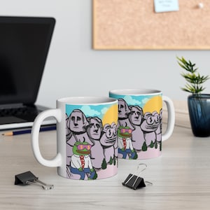 Op de afbeelding: Twee witte keramische mokken met een humoristisch ontwerp. Het ontwerp toont een cartoonkikker met een zonnebril en een pak, geplaatst op een achtergrond in Mount Rushmore-stijl. De achtergrond omvat een gele zon, blauwe lucht en groene bomen.