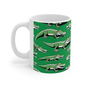 Peut inclure: Une tasse en céramique blanche avec un fond vert et un motif répété d'alligators de dessin animé. Les alligators sont verts avec des yeux jaunes et un ventre blanc.