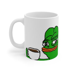 Op de afbeelding: Een witte keramische mok met een groene kikker meme die een witte mok met bruine vloeistof vasthoudt. De kikker heeft een groen lichaam, zwarte ogen en een oranje mond.