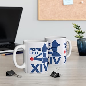 May include: Two white ceramic mugs with a blue and red graphic design. The design includes the words "POPE LEO XIV" and a stylized illustration. The mugs are on a light-colored surface.