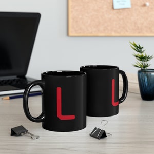 Peut inclure: Deux tasses en céramique noire avec une lettre "L" rouge sur chacune. Les tasses sont sur une surface en bois clair.