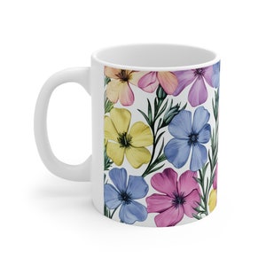 Könnte beinhalten: Weiße Keramiktasse mit einem Blumenmuster. Das Muster zeigt blaue, gelbe, rosa und hellrosa Blumen mit grünen Blättern.