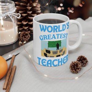 Könnte beinhalten: Eine weiße Keramiktasse mit einem blau-weißen Design. Die Tasse trägt die Aufschrift "World's Greatest Teacher" und zeigt ein Cartoon-Bild eines Lehrers, der vor einer Tafel steht.