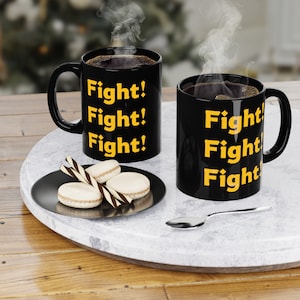 Peut inclure: Deux tasses à café noires avec le mot "Fight!" répété sur chaque tasse en lettres jaunes. Les tasses sont sur une surface en marbre blanc avec une petite assiette de biscuits et une cuillère en argent.