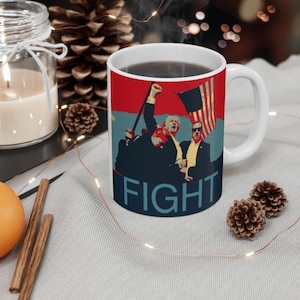 Puede incluir: Taza de café de cerámica blanca con un gráfico rojo, blanco y azul de tres personas con la palabra "FIGHT" en letras azules en la parte inferior.