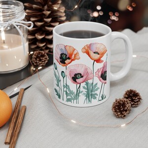Könnte beinhalten: Weiße Keramiktasse mit einem Blumendesign mit rosa und orangefarbenen Mohnblumen. Die Tasse ist mit einer dunklen Flüssigkeit gefüllt und hat einen weißen Henkel.