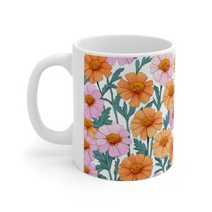Könnte beinhalten: Weiße Keramiktasse mit einem Blumenmuster. Das Muster zeigt rosa und orangefarbene Blumen mit grünen Blättern auf weißem Hintergrund.