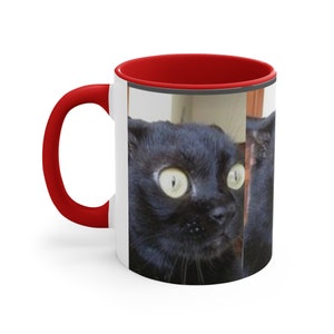 Könnte beinhalten: Eine weiße Keramiktasse mit rotem Rand und einem schwarzen Kater mit großen Augen, der darauf gedruckt ist. Der Kater schaut nach rechts im Bild.
