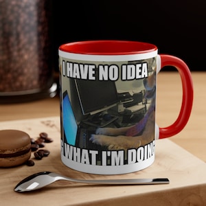 Puede incluir: Taza de café de cerámica blanca con borde y asa rojos. La taza presenta una imagen de meme de un perro sentado frente a un ordenador con el texto "No tengo ni idea de lo que estoy haciendo".