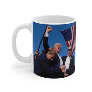 May include: A white ceramic mug with a photo of a man in a suit raising his fist in the air. The man is surrounded by other people, including a woman in a black suit. The background is a blue sky with a large American flag in the upper right corner.