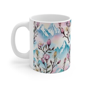 Könnte beinhalten: Weiße Keramiktasse mit einem farbenfrohen Design mit rosa Magnolienblüten, grünen Blättern und blauen Bergen. Der Hintergrund ist ein Aquarell in Rosa, Blau und Gelb.