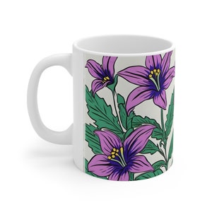 Könnte beinhalten: Weiße Keramiktasse mit einem Blumendesign. Das Design zeigt lila Blumen mit gelben Zentren und grünen Blättern auf cremefarbenem Hintergrund.