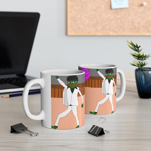 Może przedstawiać: Dwa białe ceramiczne kubki z zieloną postacią żaby w białym garniturze, która tańczy. Jeden kubek ma za żabą fioletowe i białe kółko, drugi różowe i białe kółko.