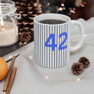 Könnte beinhalten: Eine weiße Keramiktasse mit schwarzen Streifen und der Zahl 42 in Blau. Die Tasse ist mit einer dunklen Flüssigkeit gefüllt und Dampf steigt aus der Oberseite auf.