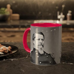 Könnte beinhalten: Eine rote und weiße Keramiktasse mit einem Schwarzweißbild eines bärtigen Mannes in Militäruniform. Die Tasse hat einen roten Rand und einen roten Griff.