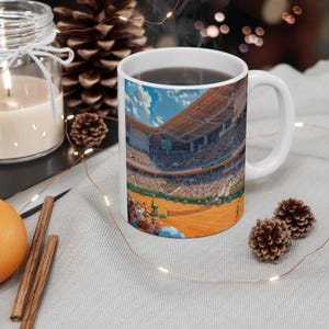 Könnte beinhalten: Weiße Keramiktasse mit einem bunten Bild eines Tennisplatzes und Zuschauern. Die Tasse ist mit einer dunklen Flüssigkeit gefüllt, wahrscheinlich Kaffee oder Tee.