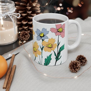 Peut inclure: Une tasse en céramique blanche avec un motif floral composé de fleurs roses, jaunes et bleues. La tasse est remplie de café noir et de la vapeur s'élève du haut.