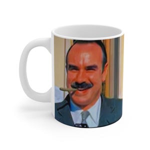 Könnte beinhalten: Weiße Keramiktasse mit einem Farbbild eines Mannes mit Schnurrbart und einer Zigarre im Mund. Der Mann trägt einen dunkelblauen Anzug und ein weißes Hemd. Der Hintergrund ist ein verschwommenes Bild eines Raumes.