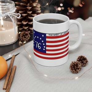 Könnte beinhalten: Weiße Keramiktasse mit patriotischem Design, das die amerikanische Flagge, die Zahl "76" und den Text "THE SPIRIT OF LIBERTY" zeigt. Die Tasse ist mit einer dunklen Flüssigkeit gefüllt und hat einen weißen Henkel.