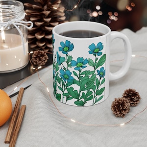 Könnte beinhalten: Eine weiße Keramiktasse mit einem blauen und grünen Blumendesign. Die Tasse ist mit einer dunklen Flüssigkeit gefüllt und hat einen Henkel auf der rechten Seite.