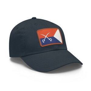 Puede incluir: Una gorra de béisbol azul marino con un parche que muestra una bandera roja, blanca y azul con espadas cruzadas.