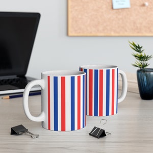Könnte beinhalten: Zwei weiße Keramikbecher mit roten und blauen vertikalen Streifen. Die Becher stehen auf einem weißen Tisch mit einem Bleistift und Büroklammern.