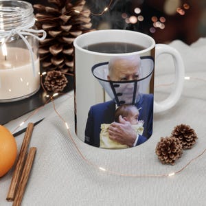 May include: White ceramic coffee mug with a printed image of a man wearing a cone around his neck holding a baby. The man is wearing a dark suit and a white shirt. The baby is wearing a yellow shirt.
